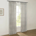 A pair of pleated white curtains with a subtle dark leaf pattern hangs from a black rod in a bright room with a window showing green trees outside and a framed botanical print on the wall
