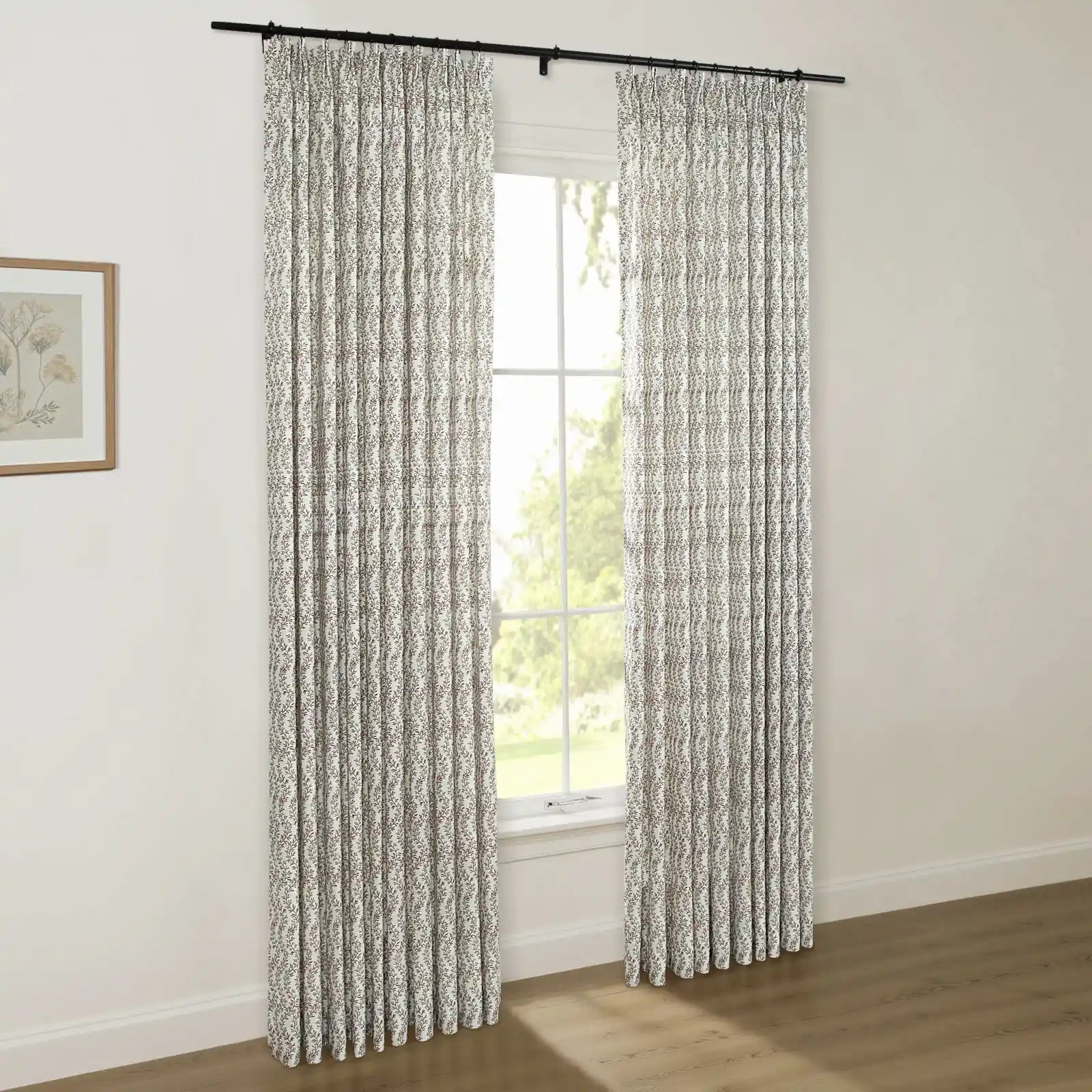 A pair of pleated white curtains with a subtle dark leaf pattern hangs from a black rod in a bright room with a window showing green trees outside and a framed botanical print on the wall
