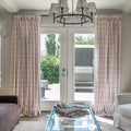 A modern living room features elegant cream botanical print curtains framing glass patio doors, a contemporary black chandelier with white drum shades, and a glass coffee table topped with fabric samples, looking out onto a bright green patio