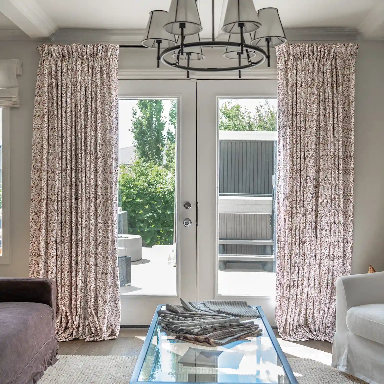 A modern living room features elegant cream botanical print curtains framing glass patio doors, a contemporary black chandelier with white drum shades, and a glass coffee table topped with fabric samples, looking out onto a bright green patio