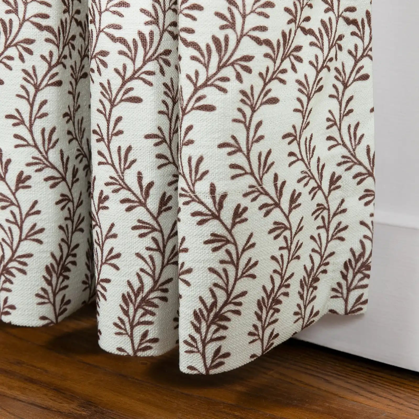 Textile print of a white textured curtain with a repeating brown stylized leaf pattern draped over a rich dark wooden floor next to a white baseboard