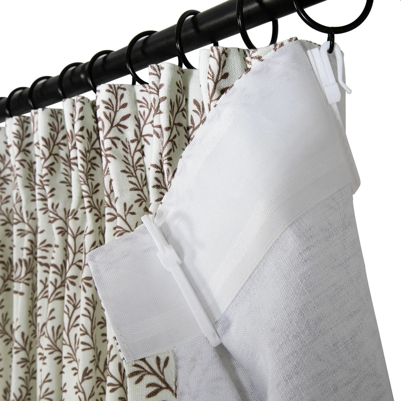 Close-up of pleated white fabric curtains featuring a brown branch-leaf pattern hanging on a black rod with rings, detailed with a white textured lining panel and white plastic pin hooks