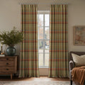 A modern living room featuring window curtains with a green, orange, and red plaid pattern framing a view of trees and grass, beside a wooden dresser with a blue-green vase of foliage and a grey armchair with an orange throw