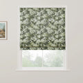 A fabric Roman blind featuring an intricate green and white floral pattern is partially raised in a white window frame revealing blurred green trees outside, with a rustic landscape painting on the adjacent white wall