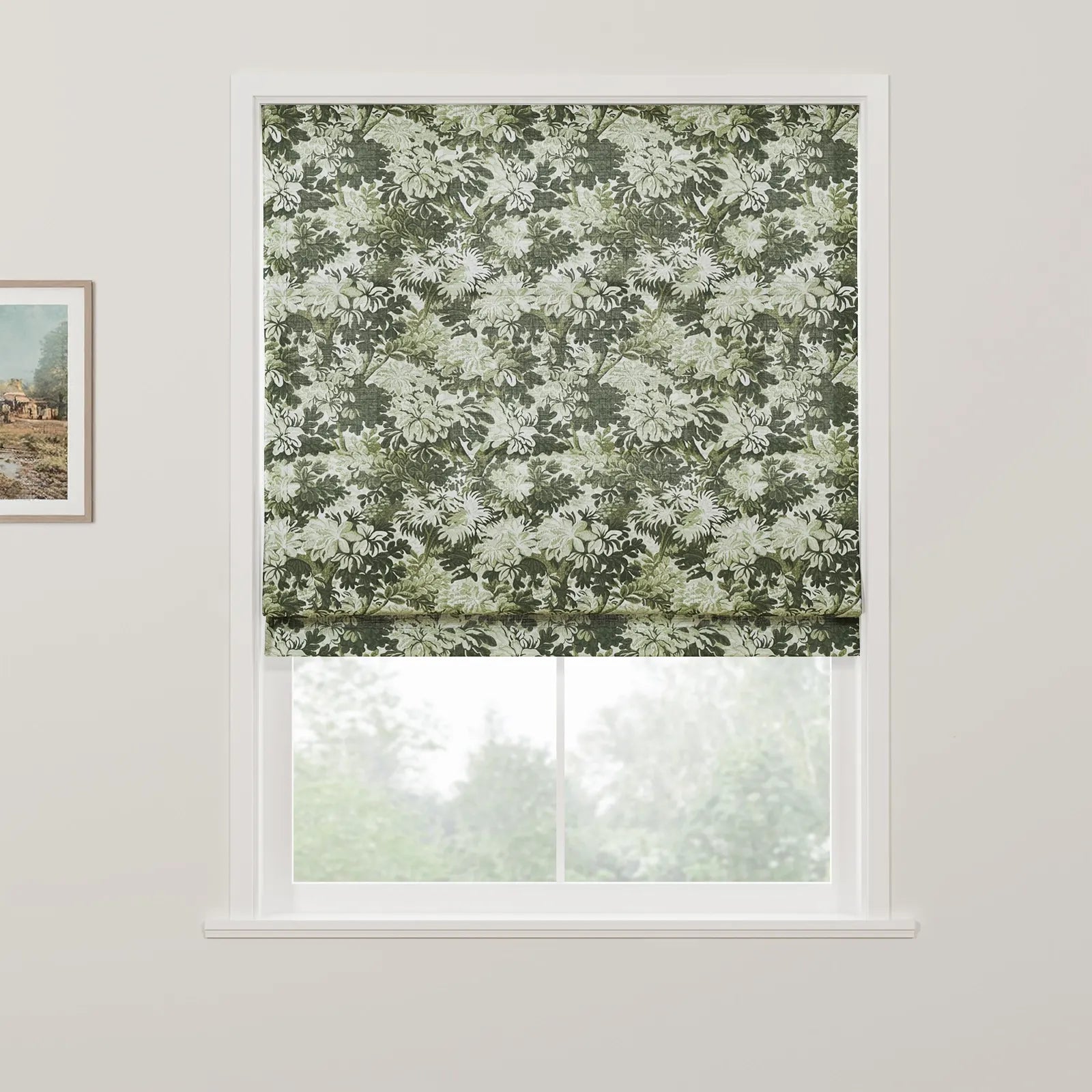 A fabric Roman blind featuring an intricate green and white floral pattern is partially raised in a white window frame revealing blurred green trees outside, with a rustic landscape painting on the adjacent white wall