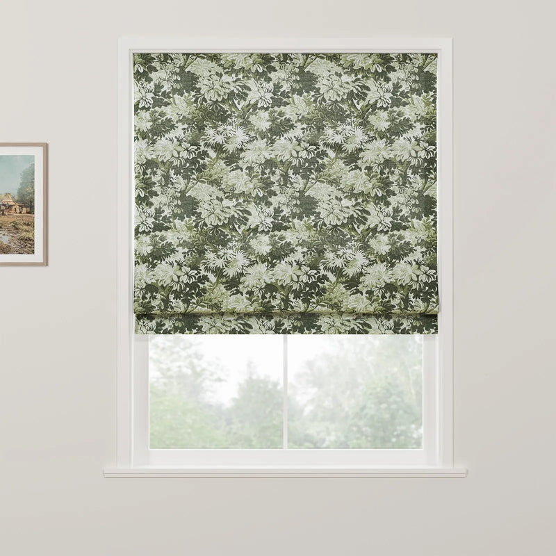 A fabric Roman blind featuring an intricate green and white floral pattern is partially raised in a white window frame revealing blurred green trees outside, with a rustic landscape painting on the adjacent white wall