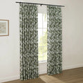A pair of pleated green and white floral curtains hang from a black rod in a bright room, framing a window with a view of outdoor greenery, beside a framed botanical print on a white wall above light wooden flooring