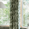 A green and white intricate floral curtain partially covers a white grid-paned window offering a view of vibrant green outdoor foliage and tree branches