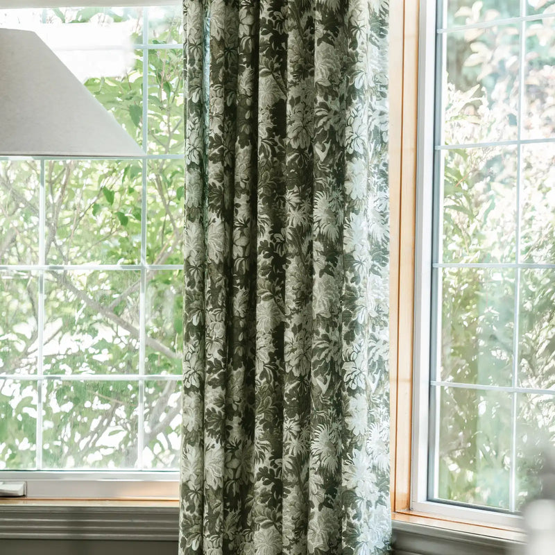 A green and white intricate floral curtain partially covers a white grid-paned window offering a view of vibrant green outdoor foliage and tree branches