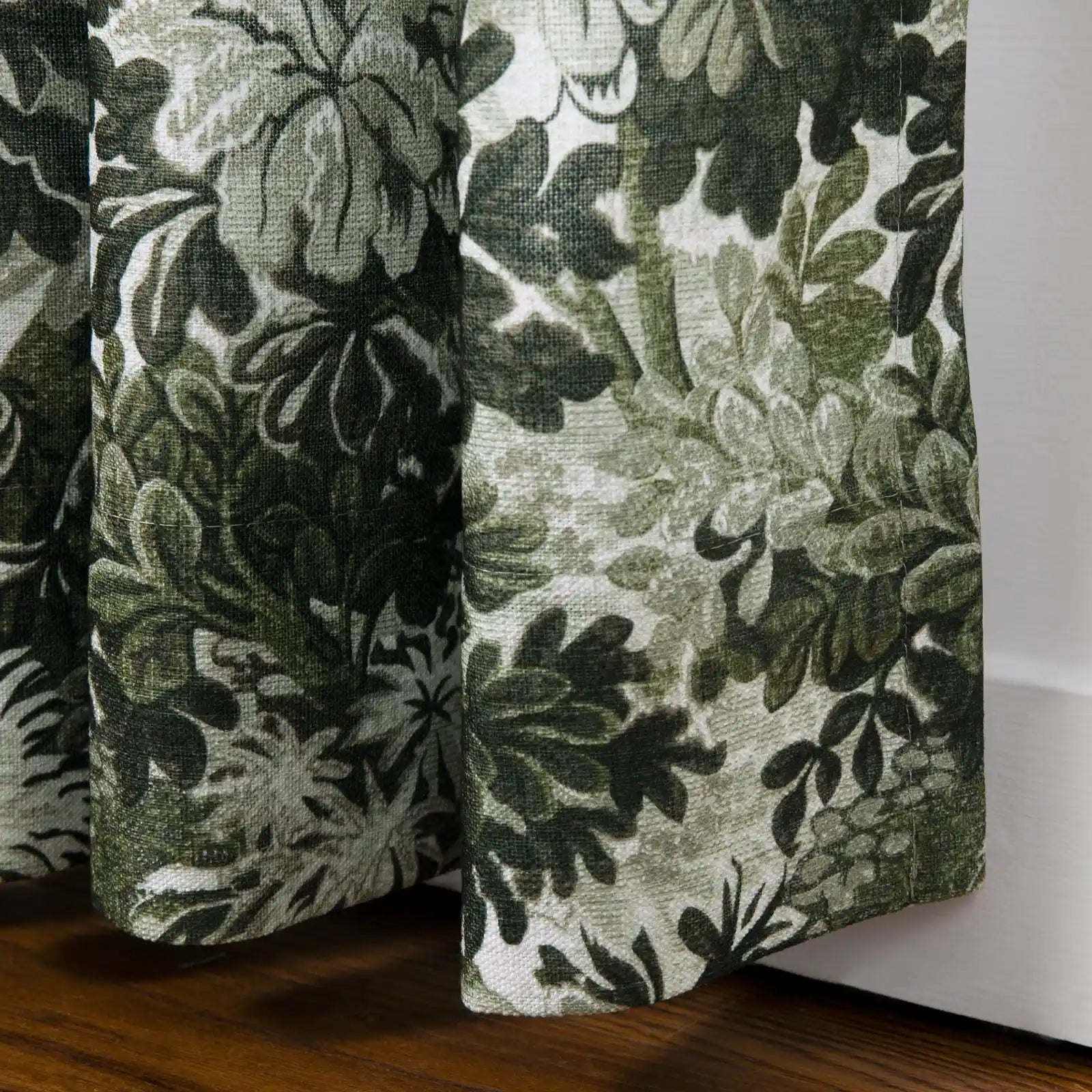 Photograph of fabric panel featuring a dark green and light green botanical pattern of leaves and flowers on a textured white background, draped over a wooden floor