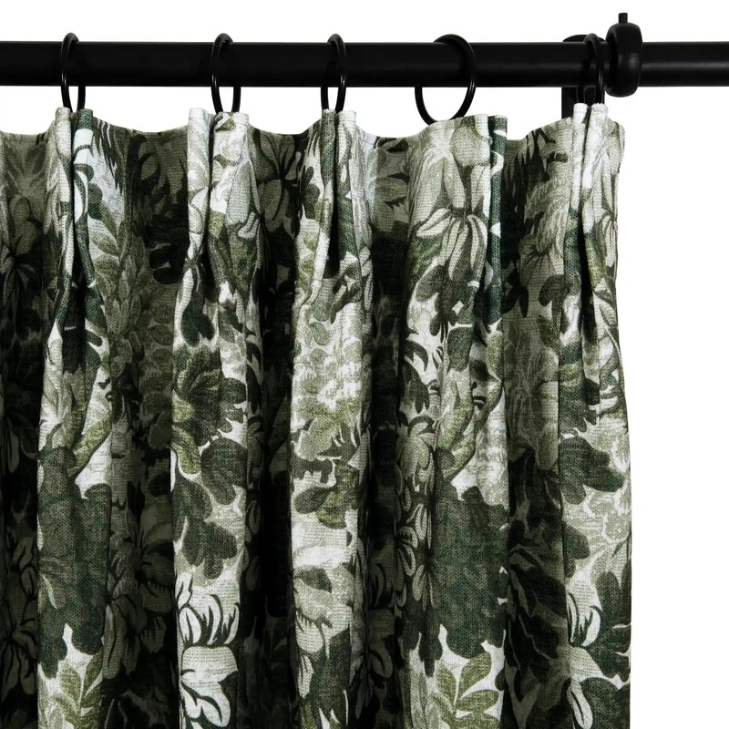 Textile curtain displaying a dense dark and light green botanical pattern with pleated fabric hanging from a black curtain rod and rings
