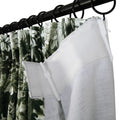 Close-up of pleated green and white tropical leaf patterned curtains hanging on a black metal rod with rings, revealing a white textured privacy lining with plastic hooks