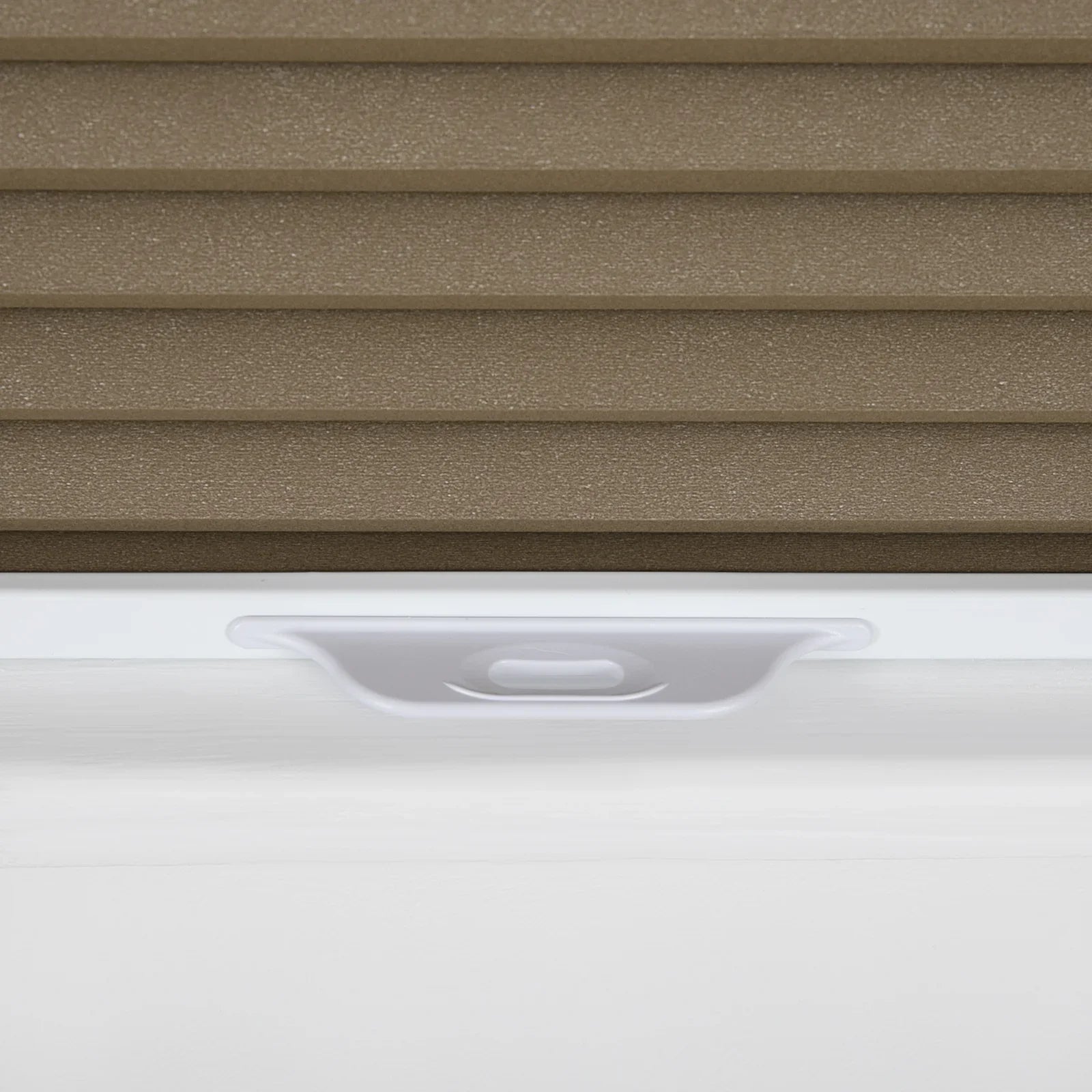 Close-up of a brown textured pleated cellular window Blind with a white bottom rail and an integrated white pull handle resting on a white window sill