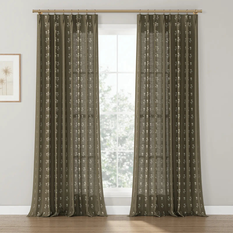 Dark olive-brown textured window curtains with a repeating white floral block print pattern, suspended from a brass rod, illuminating a room with light hardwood floors and a framed palm tree art print on the left wall