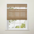 A natural tan woven bamboo blind is partially raised on a white multi-pane window, revealing blurred green trees outside and featuring a small metal ring