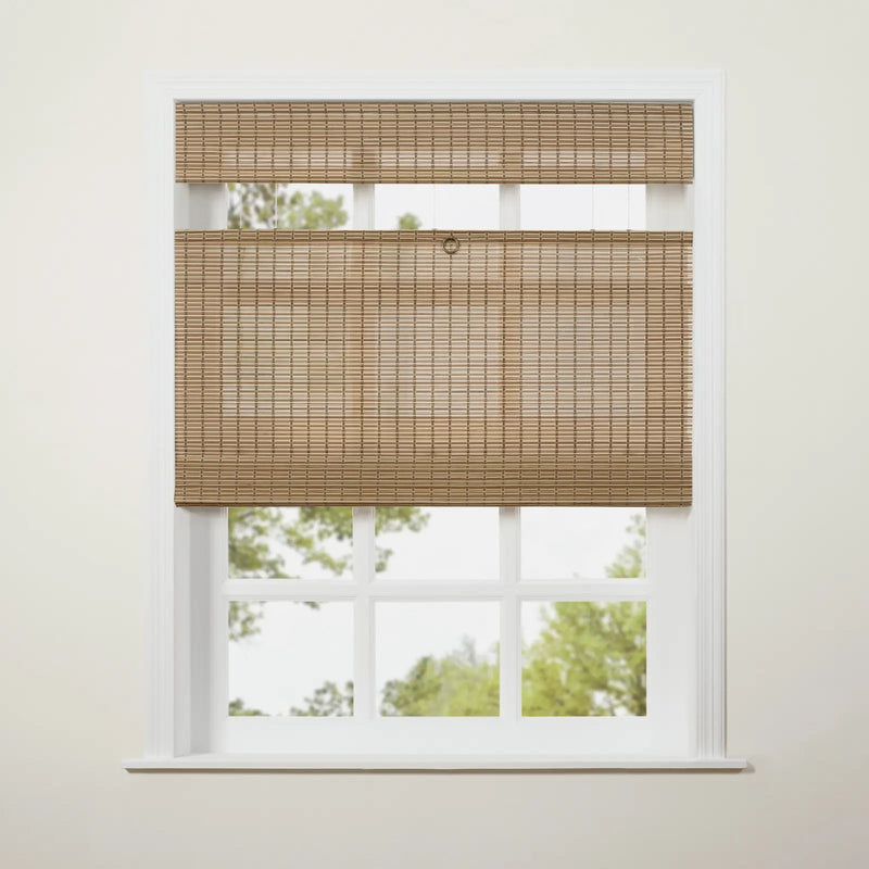 A natural tan woven bamboo blind is partially raised on a white multi-pane window, revealing blurred green trees outside and featuring a small metal ring