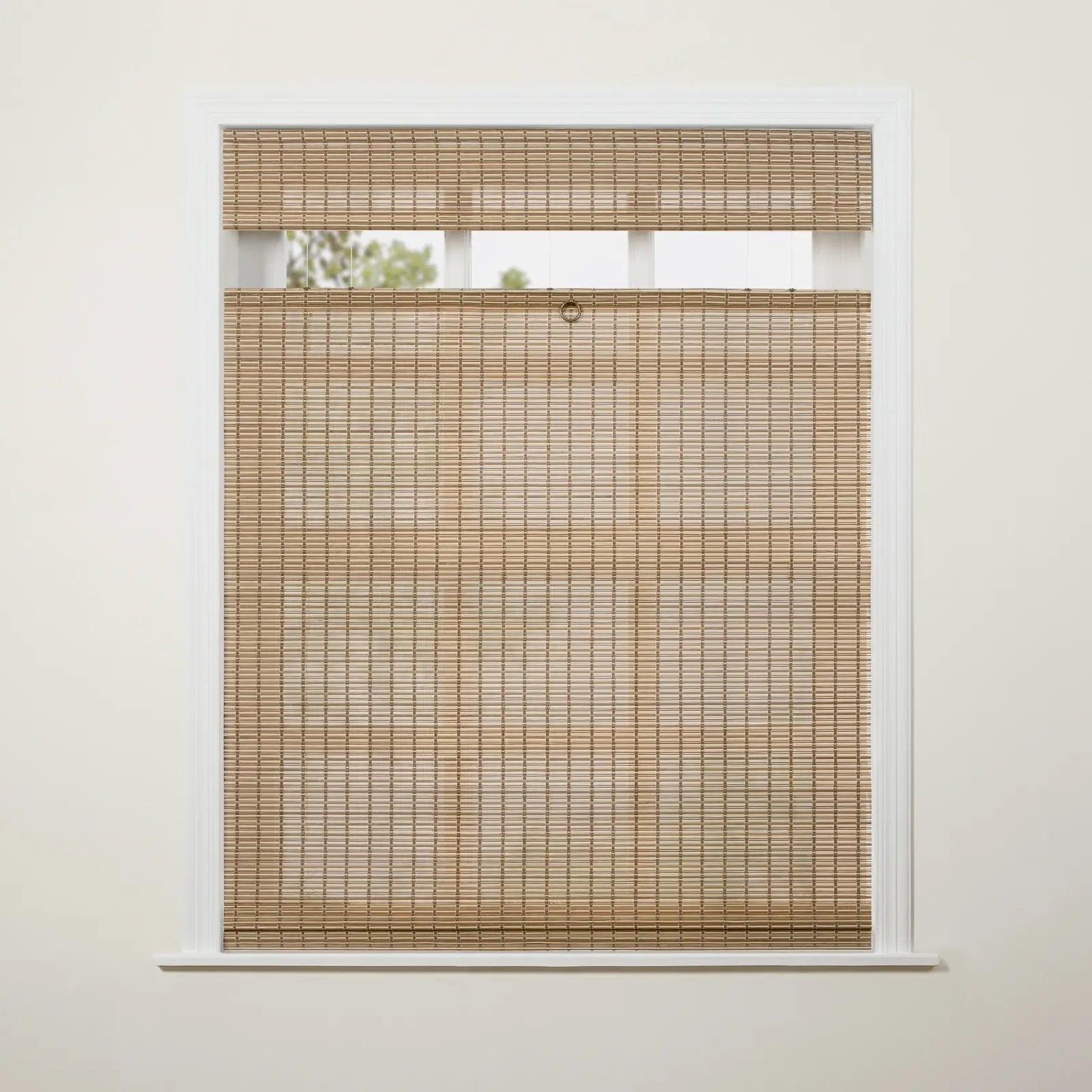 Photograph of a white framed window featuring natural light brown woven bamboo blinds in a top-down/bottom-up style with a brass ring pull, revealing blurred green foliage through the upper opening