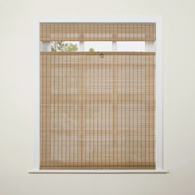 Photograph of a white framed window featuring natural light brown woven bamboo blinds in a top-down/bottom-up style with a brass ring pull, revealing blurred green foliage through the upper opening