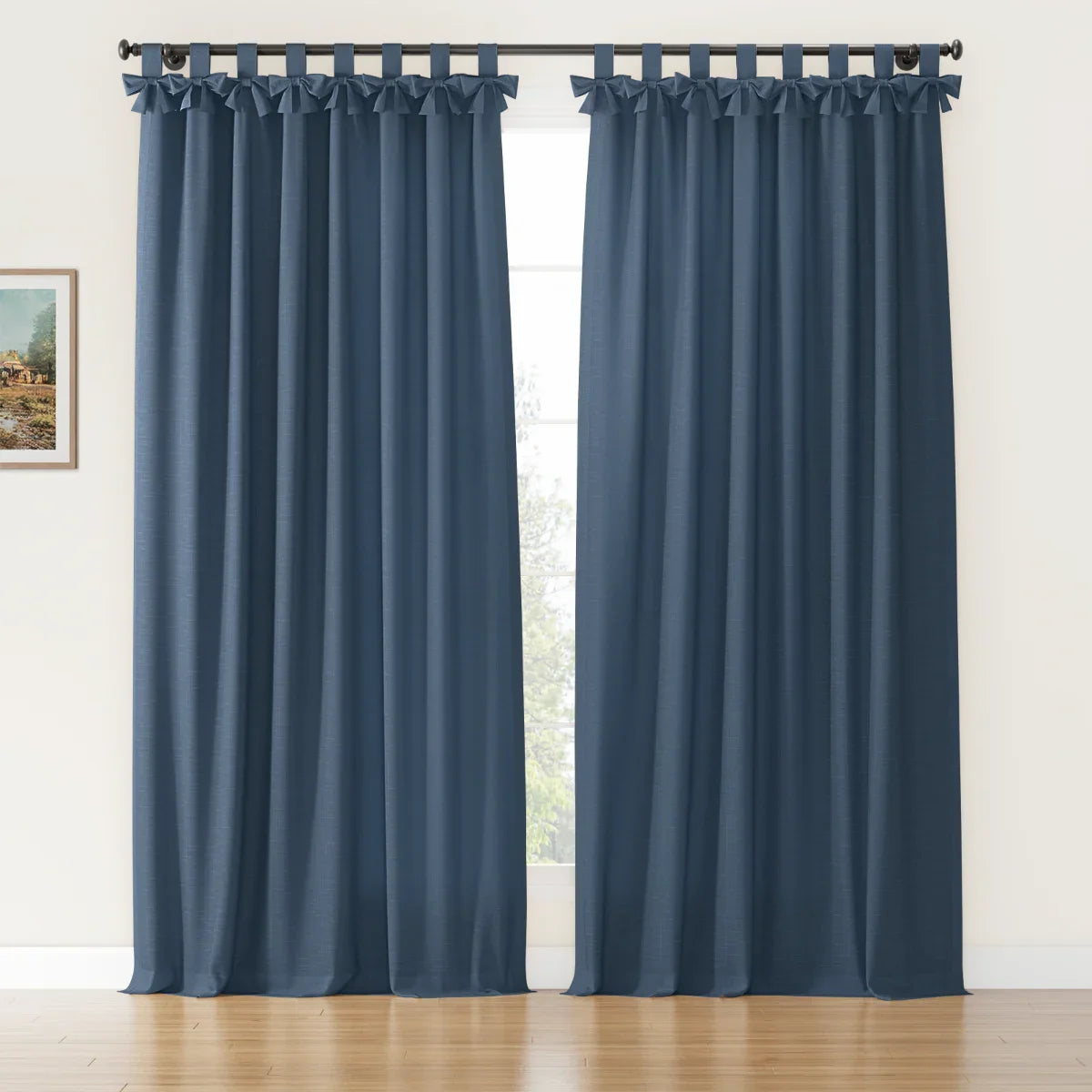 A pair of textured indigo blue tab top curtains featuring decorative bow ties hangs on a black rod in a bright room with a polished hardwood floor and a framed landscape painting on the left wall