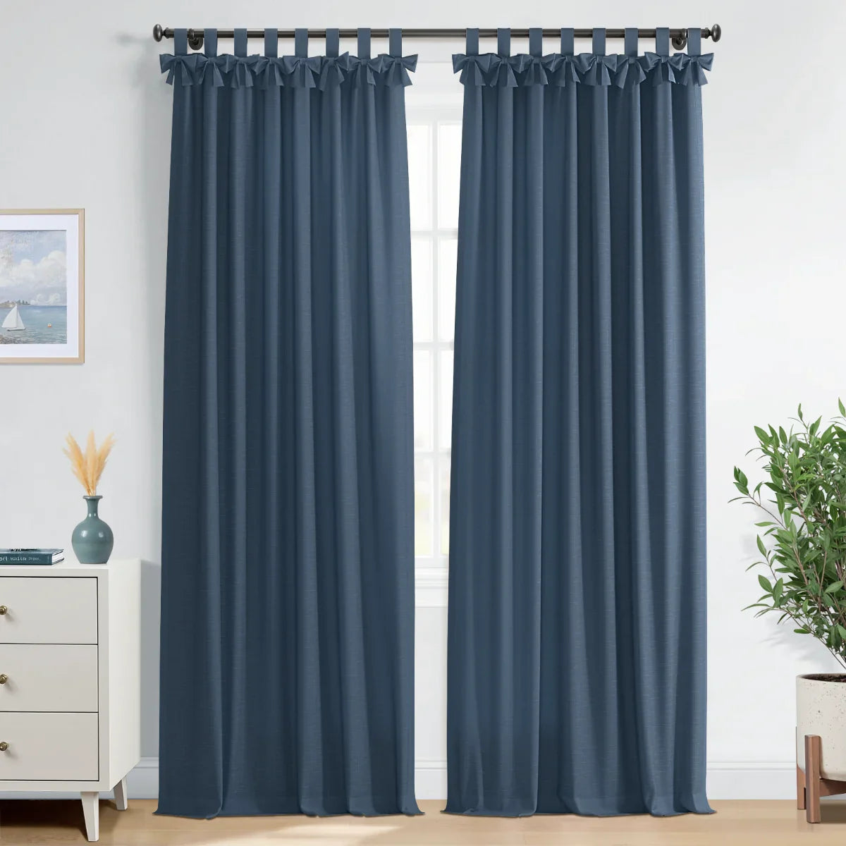 Textured dark blue tab top curtains with decorative bows frame a window in a bright room, featuring a white dresser with a teal vase and dried pampas grass, a sailboat artwork, and a potted green plant