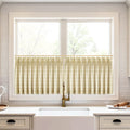 Photograph of a bright modern kitchen with beige plaid cafe curtains on a window above a white farmhouse sink, featuring an antique brass faucet with a textured handle and white subway tile backsplash