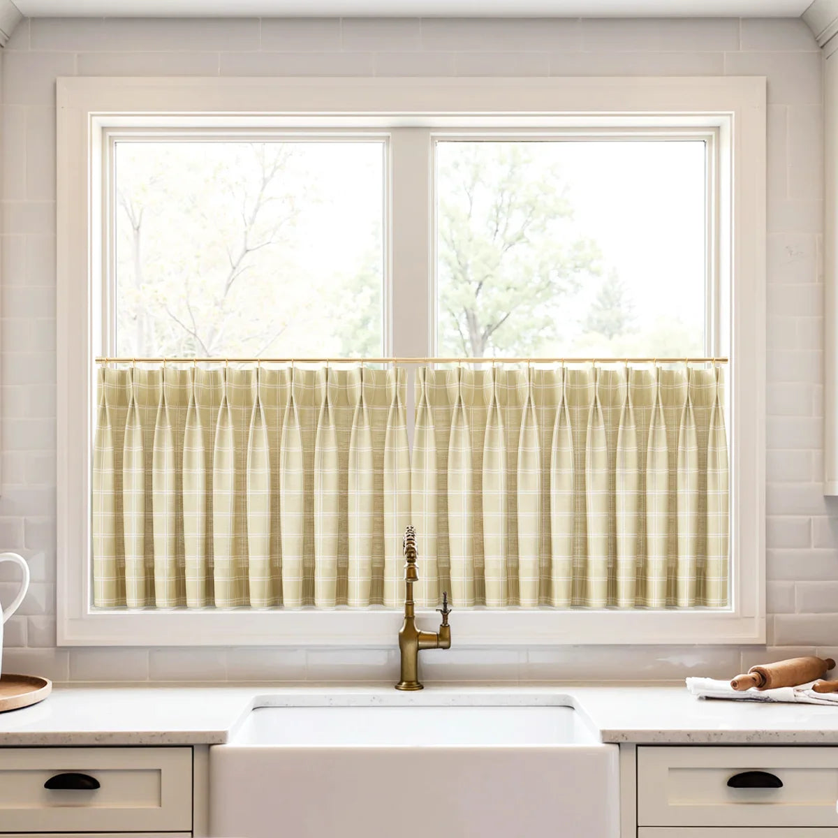 Photograph of a bright modern kitchen with beige plaid cafe curtains on a window above a white farmhouse sink, featuring an antique brass faucet with a textured handle and white subway tile backsplash