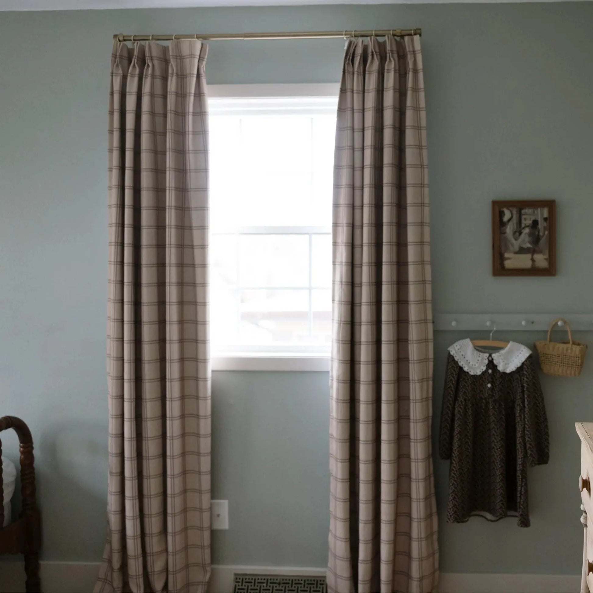 Photograph of a muted sage green room with beige plaid curtains on a brass rod, a bright window, and a child's brown dress with a white lace collar hanging next to a woven basket on a peg rail