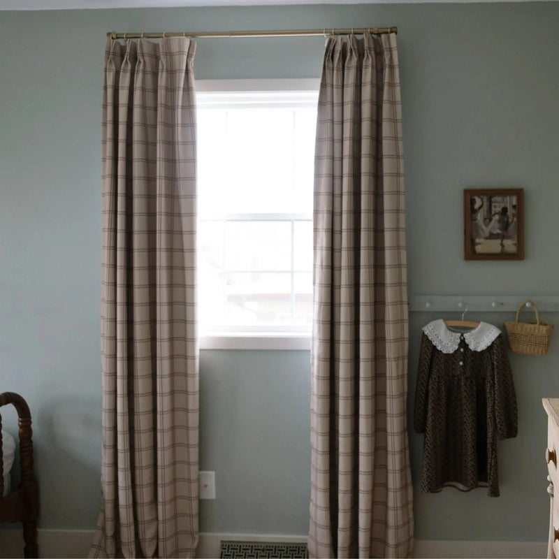 Photograph of a muted sage green room with beige plaid curtains on a brass rod, a bright window, and a child's brown dress with a white lace collar hanging next to a woven basket on a peg rail