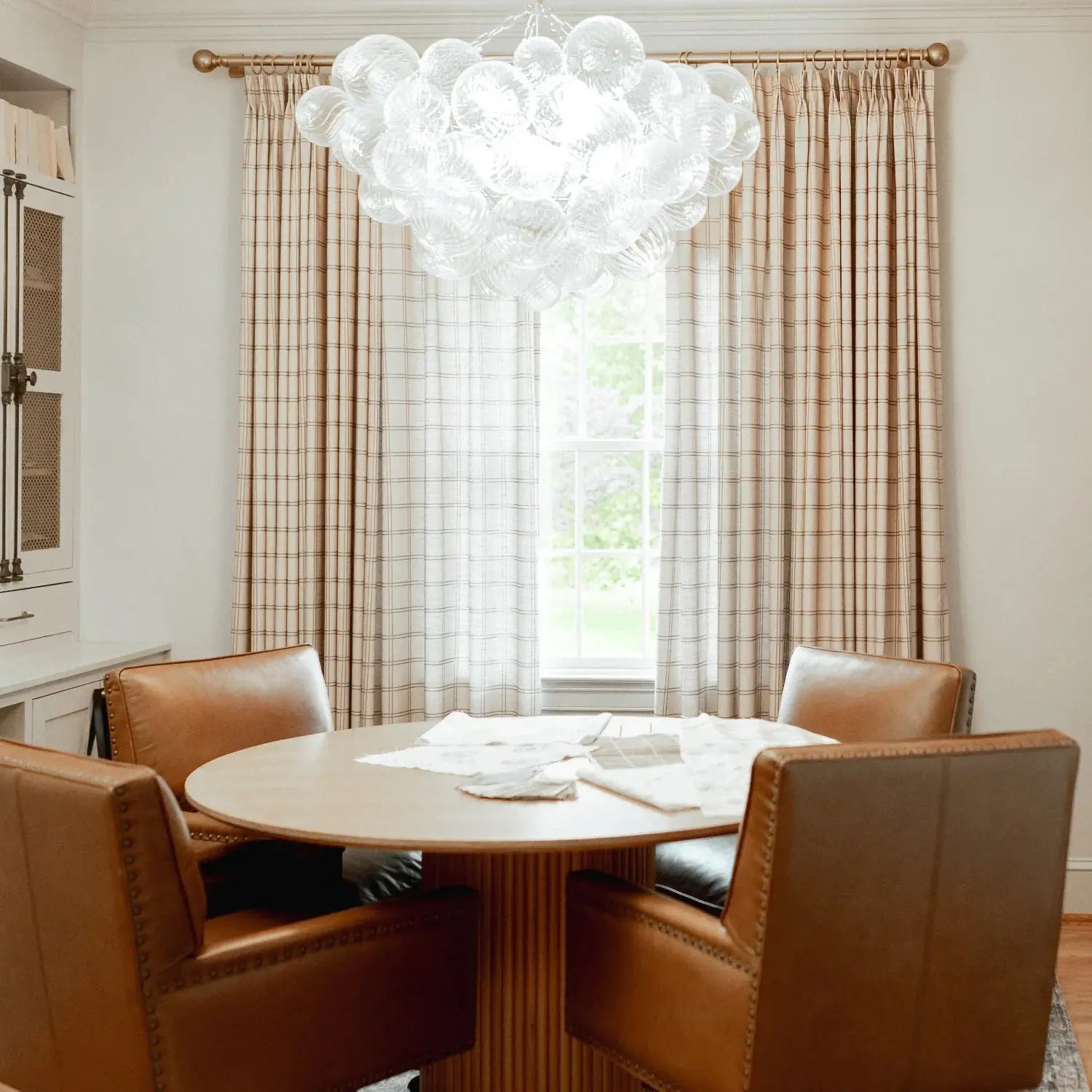 A modern dining nook featuring a large clear glass bubble chandelier a round fluted light wood table and four cognac leather chairs with brass studding set against beige plaid window curtains
