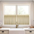 Photograph of a bright kitchen featuring a white farmhouse sink under a window with beige plaid pleated cafe curtains on a gold rod, accompanied by a vintage brass faucet and white subway tile backsplash