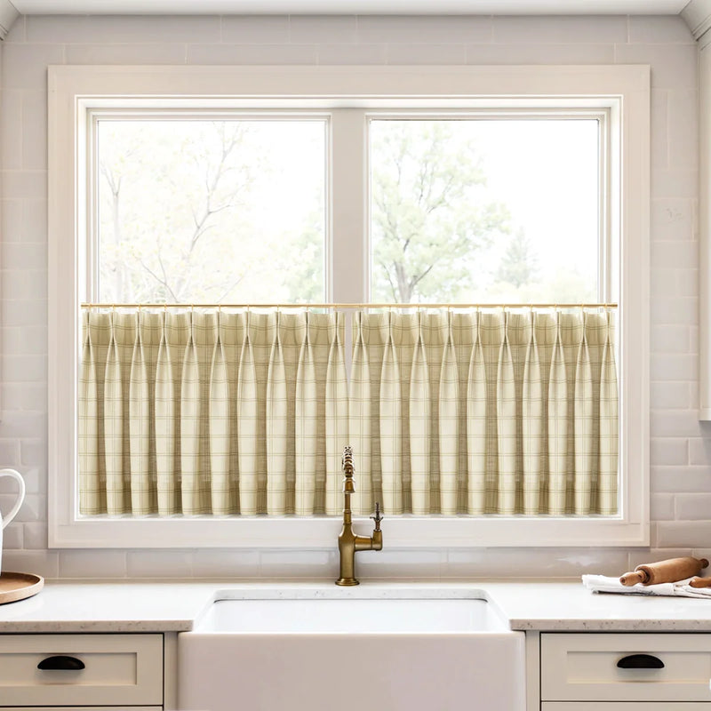 Photograph of a bright kitchen featuring a white farmhouse sink under a window with beige plaid pleated cafe curtains on a gold rod, accompanied by a vintage brass faucet and white subway tile backsplash