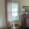 A white shaggy dog with a pink collar sits on a wooden floor in a rustic home interior next to a wicker basket filled with pinecones and string lights in front of a window with a beige plaid curtain and an antique wooden secretary desk topped with a brass lamp and a framed nautical ship painting