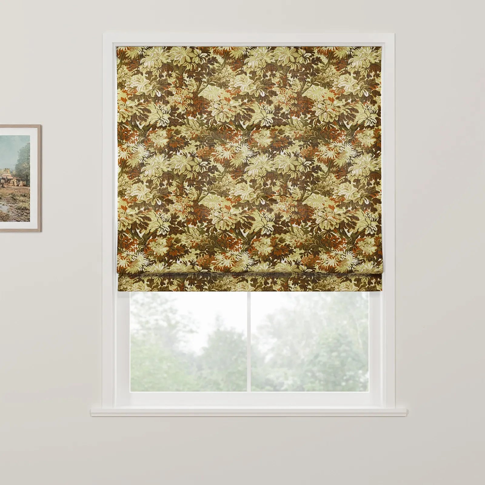 A Roman blind with a dense botanical pattern in autumn greens and browns covers the upper window, revealing blurred trees outside and a framed rustic landscape painting on the adjacent wall