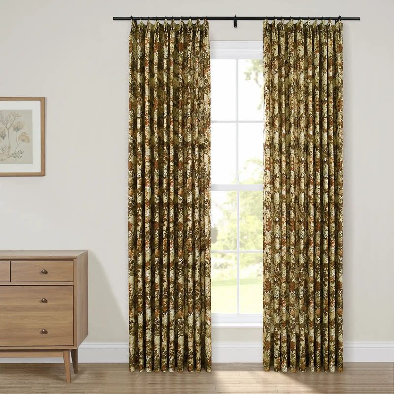 Photograph of a cozy room interior featuring pleated olive green and brown floral patterned curtains framing a window with white grid panes revealing green foliage outside, next to a mid-century modern wooden dresser and a framed botanical print on the wall