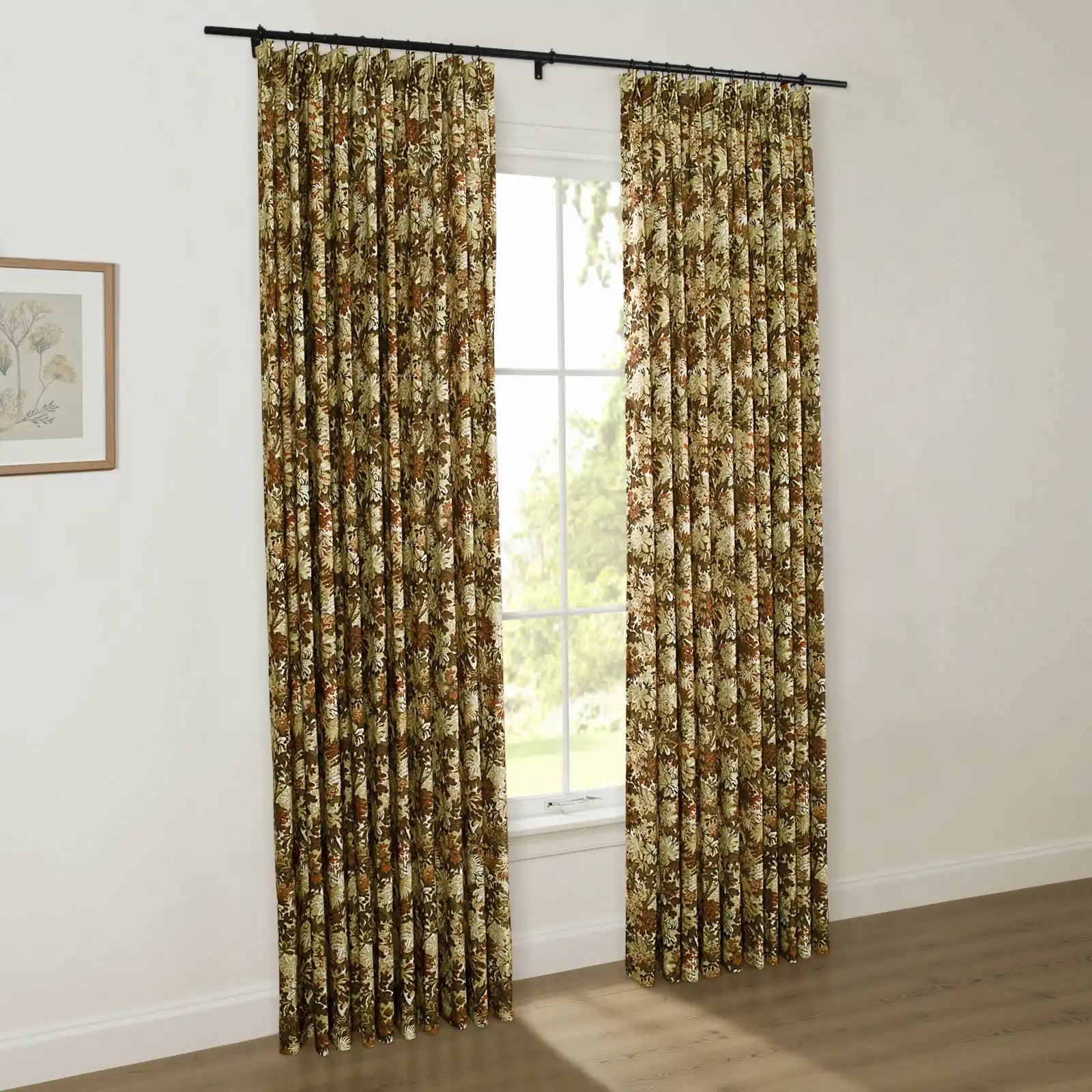 A pair of pleated earthy brown and olive green floral patterned curtains hangs from a black rod in a bright room, framing a window with a view of green trees and featuring a nearby framed botanical art print on a white wall