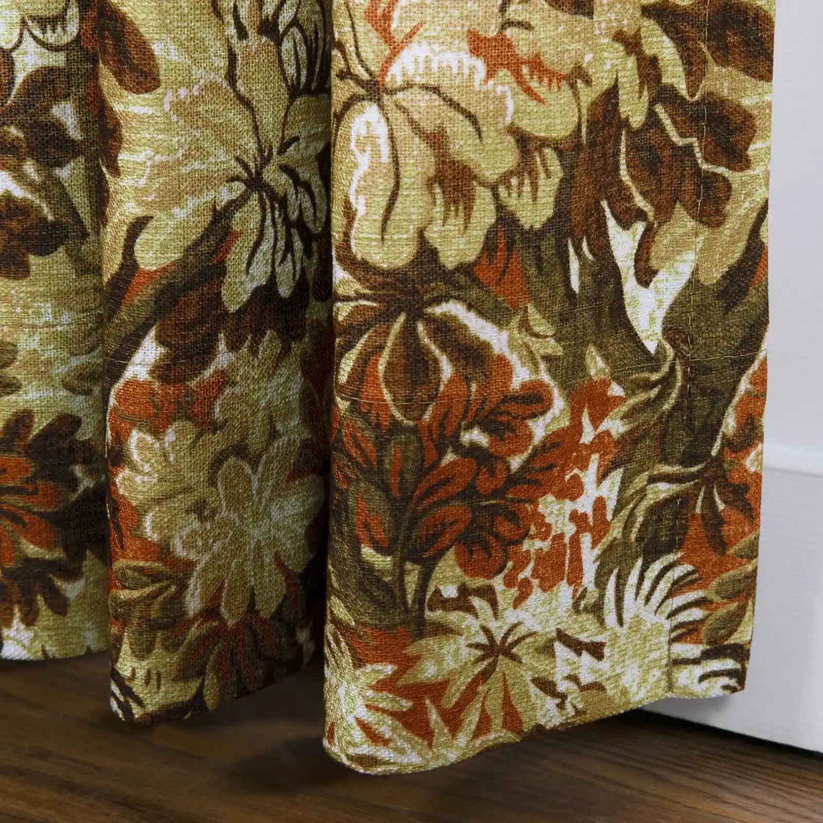 Textile curtains featuring a vintage botanical pattern of olive green, warm brown, and burnt orange rest on a dark wood floor next to a white baseboard