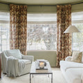 Photograph of a cozy living room featuring sage green walls, a bay window with large grid panes, floral print curtains in brown and gold, a light striped armchair draped with a blue throw, and a lit candle on a marble coffee table