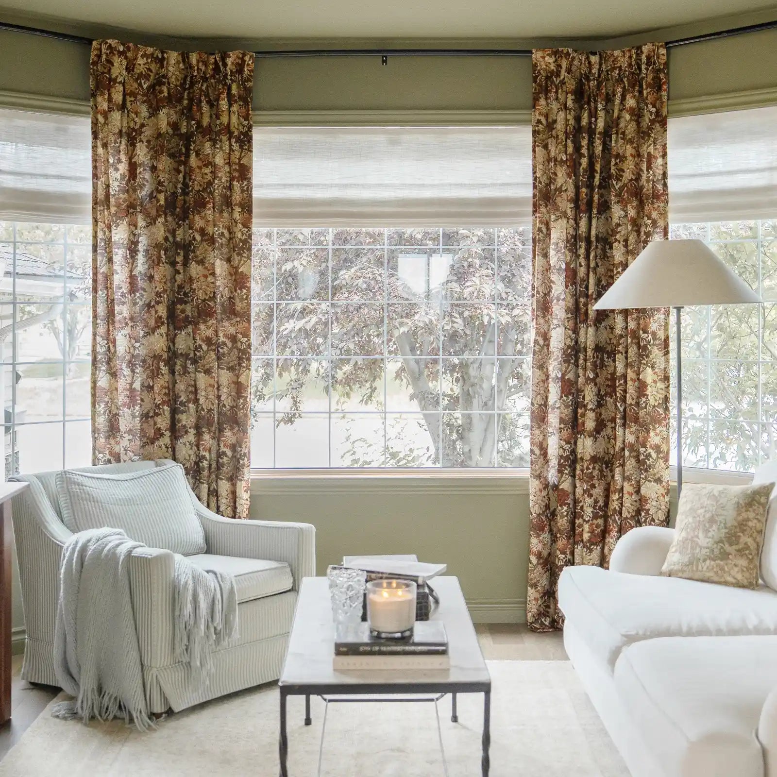 Photograph of a cozy living room featuring sage green walls, a bay window with large grid panes, floral print curtains in brown and gold, a light striped armchair draped with a blue throw, and a lit candle on a marble coffee table