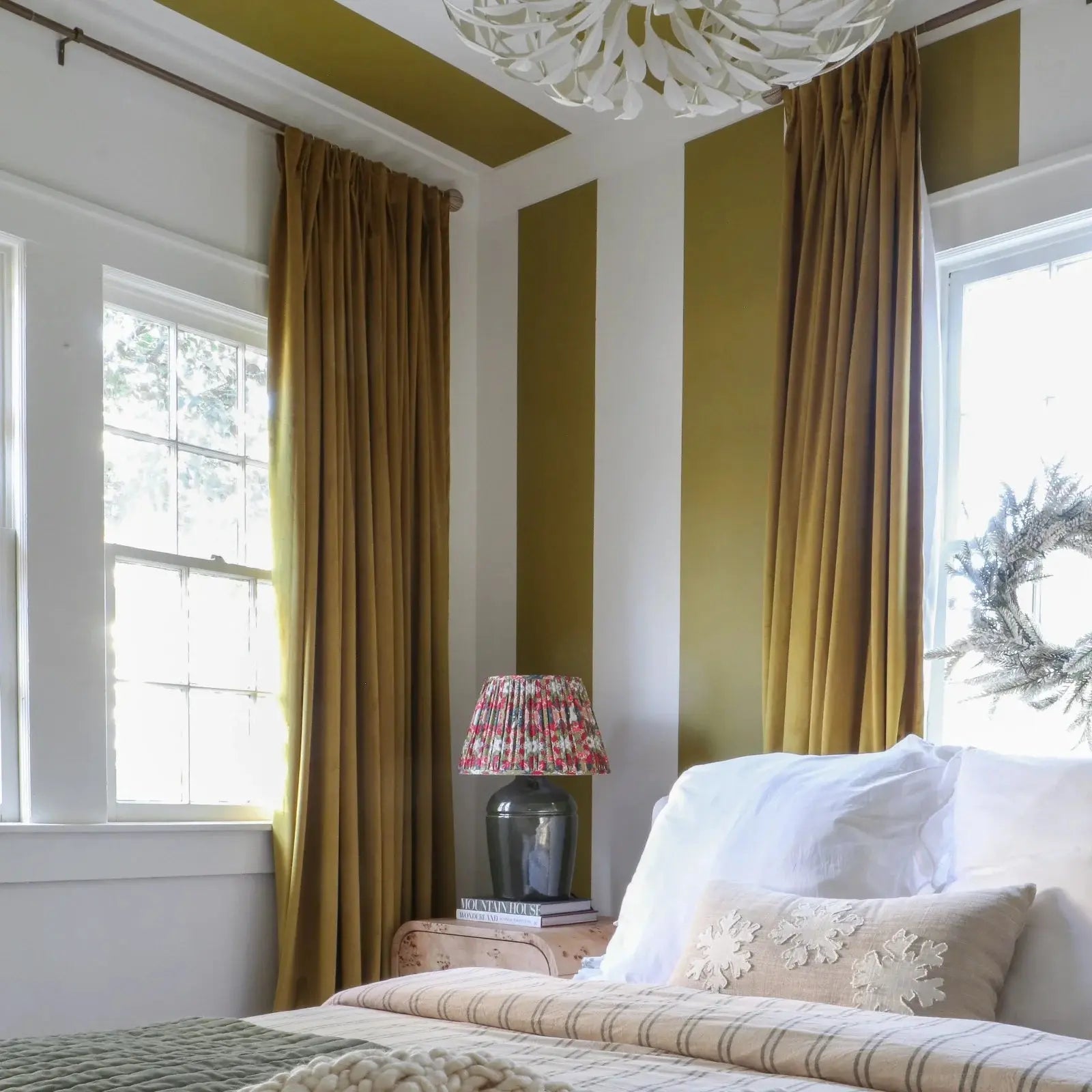 Photograph of a cozy bedroom interior with olive green and white striped walls and ceiling, mustard yellow velvet curtains framing sunlit windows, a floral pleated lampshade on a bedside table, and a bed adorned with a white snowflake pillow and a white winter wreath in the window