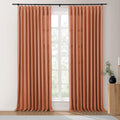 Terracotta curtains on a black rod frame a window looking onto green trees, accompanied by a fluted wooden side table, a black vase with green foliage, and a gold candlestick