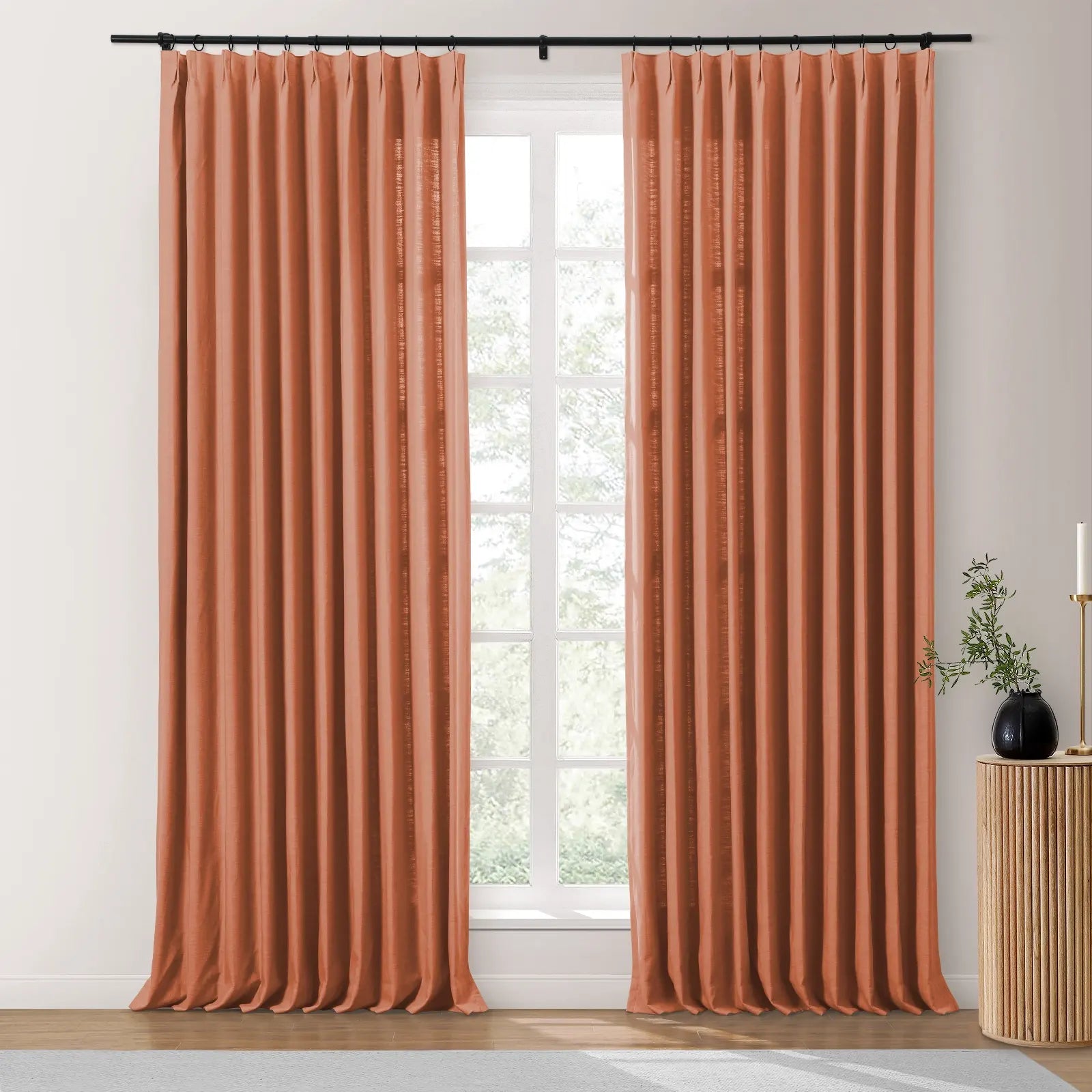 Terracotta curtains on a black rod frame a window looking onto green trees, accompanied by a fluted wooden side table, a black vase with green foliage, and a gold candlestick