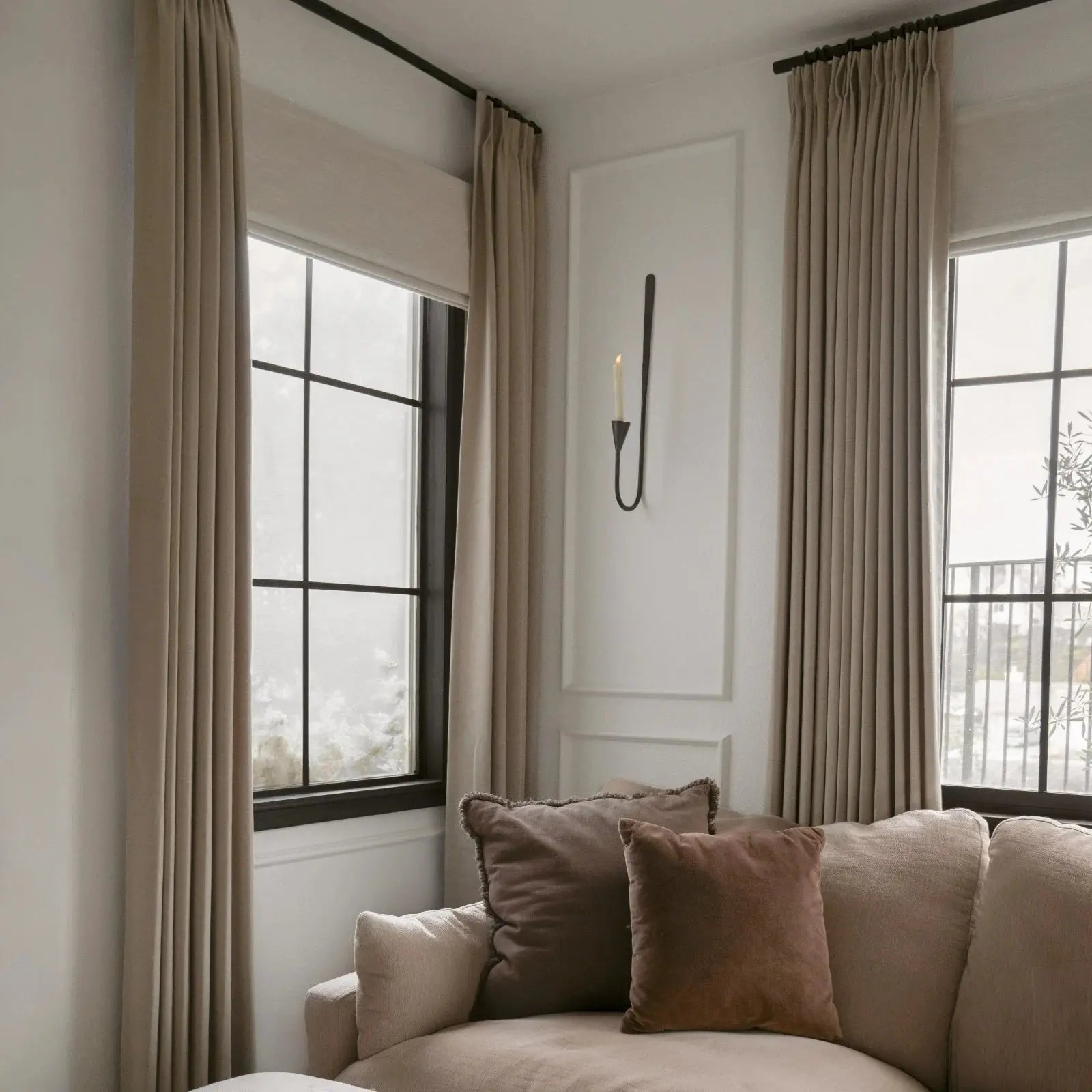 Interior of a cozy living room featuring a neutral-toned sectional sofa with plush dark brown velvet pillows, flowing beige linen curtains, and a sleek black wall sconce with a lit candle on a white paneled wall