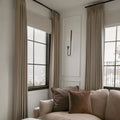 Interior of a cozy living room featuring a neutral-toned sectional sofa with plush dark brown velvet pillows, flowing beige linen curtains, and a sleek black wall sconce with a lit candle on a white paneled wall