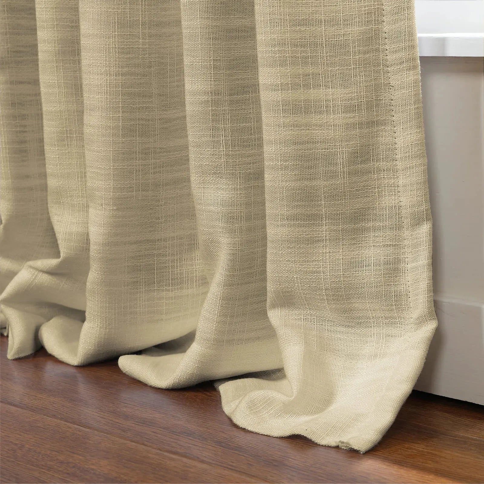 Photograph of cream linen-blend curtains with a visible woven texture, softly draped in elegant folds and puddling on a rich brown hardwood floor beside a white window sill