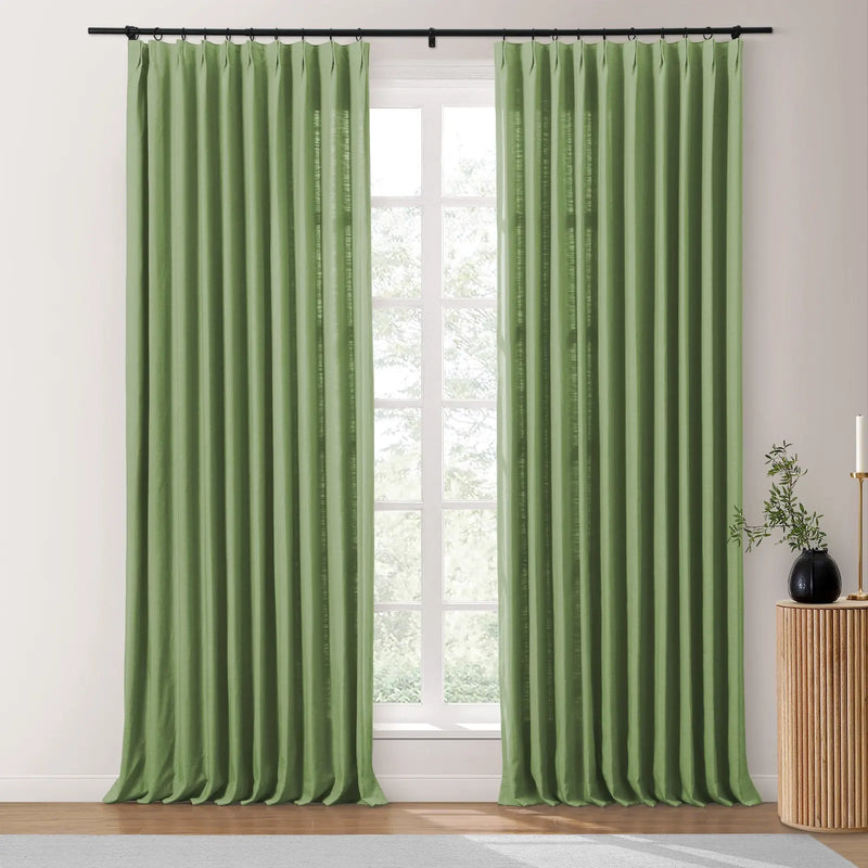 Sage green linen-look pinch-pleat curtains on a black rod covering a multi-paned window, next to a fluted wooden table with a black vase and candle holder