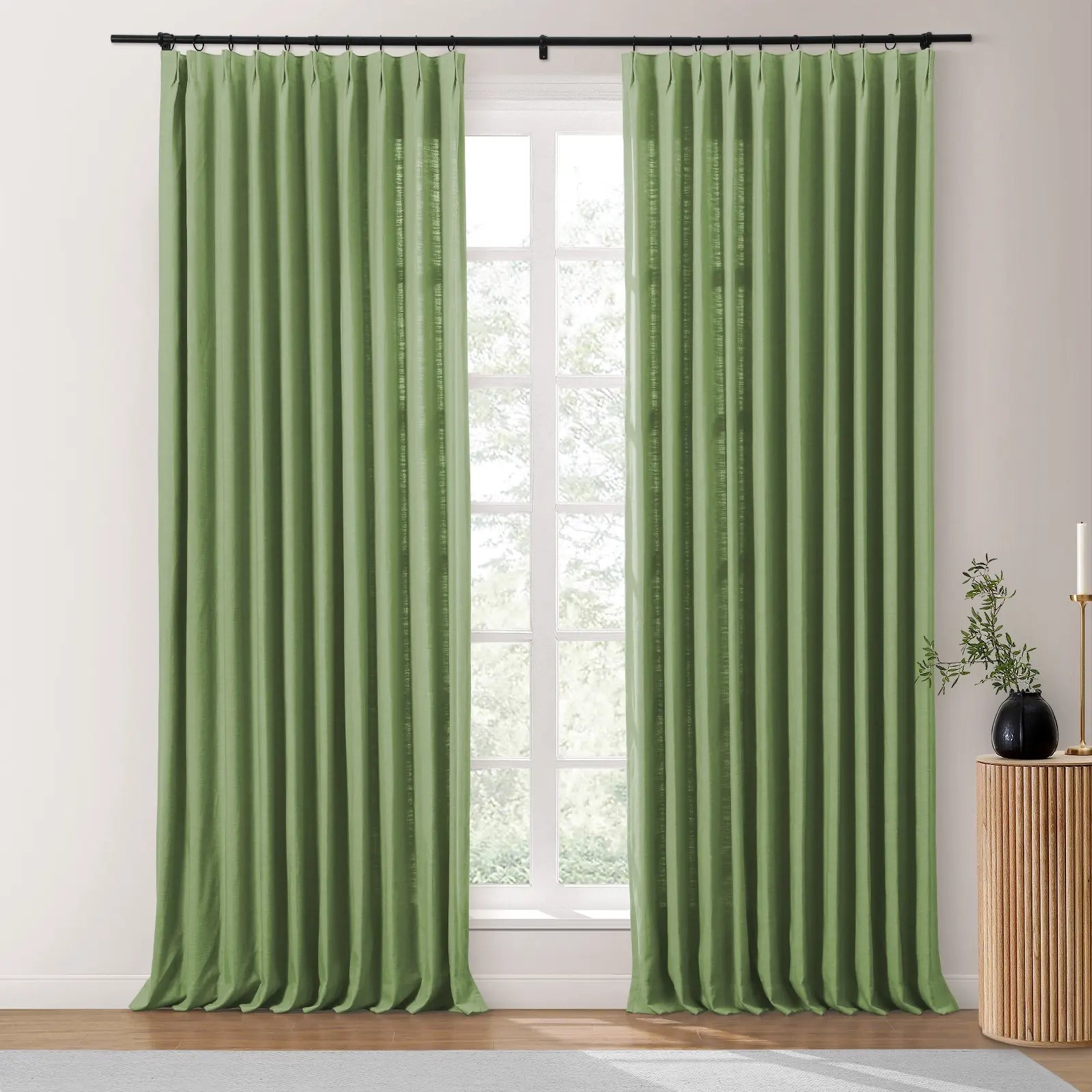 Sage green linen-look pinch-pleat curtains on a black rod covering a multi-paned window, next to a fluted wooden table with a black vase and candle holder