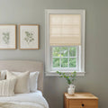 A serene bedroom interior features a window with a beige roman shade looking out to lush green trees, a bed with neutral textured pillows, two framed botanical prints above the headboard, and a wooden nightstand with a white ribbed vase holding fresh greenery