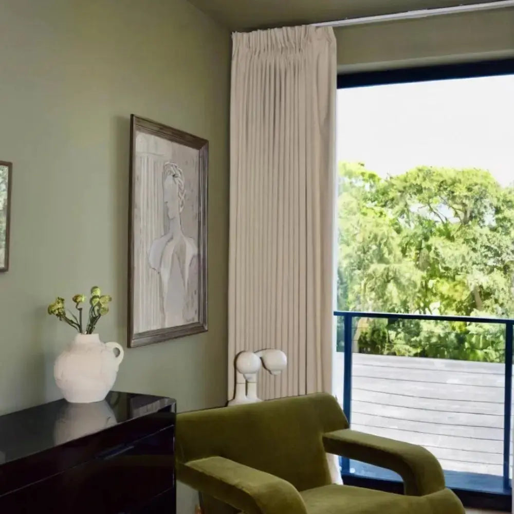 Photograph of a modern interior room with olive green walls, a matching velvet armchair, a glossy black dresser holding a white vase with lime green flowers, an abstract framed bust artwork, and a large window with cream curtains showing lush green trees outside
