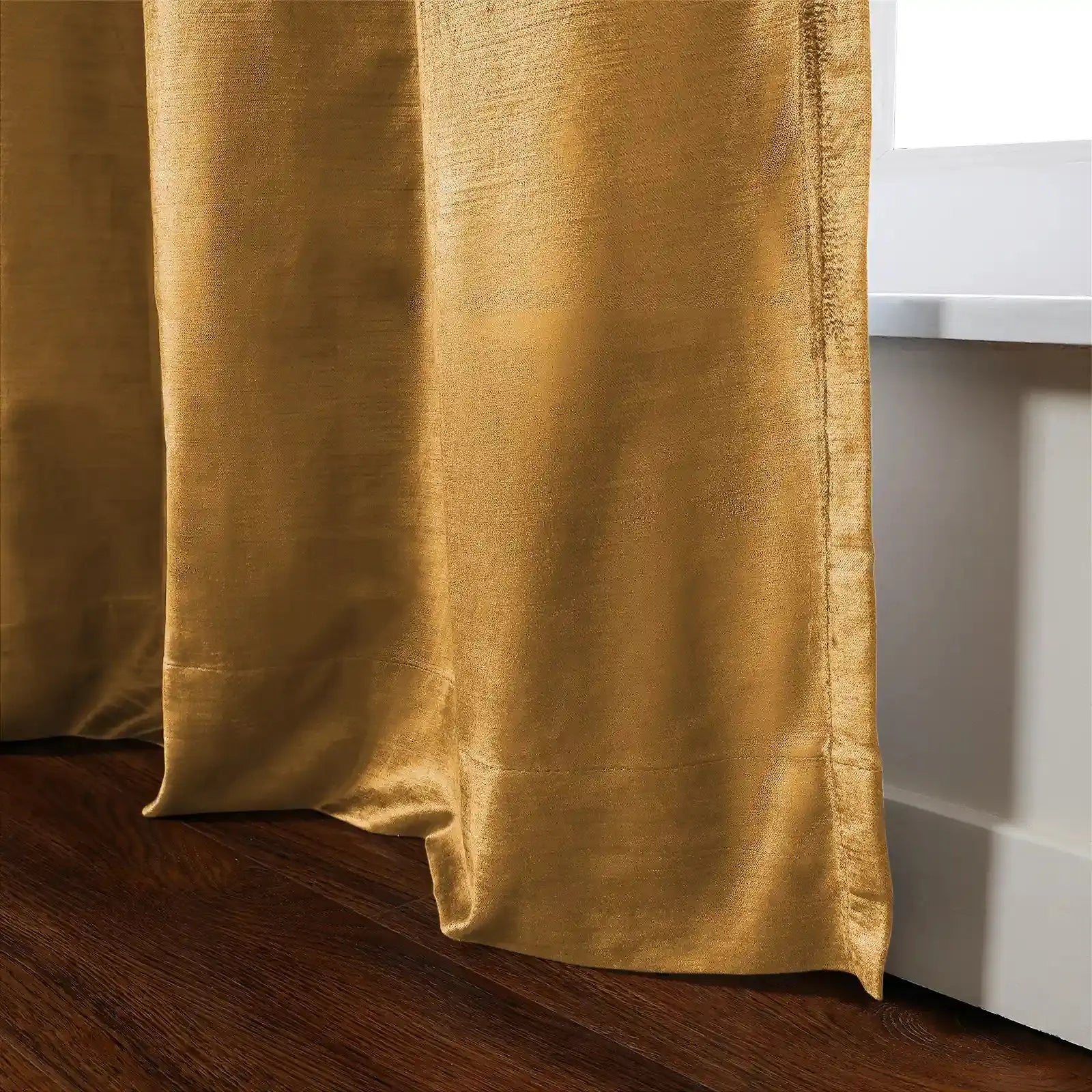 Golden yellow curtain of lustrous slub-textured fabric with cascading folds resting on a dark wood floor next to a white window frame