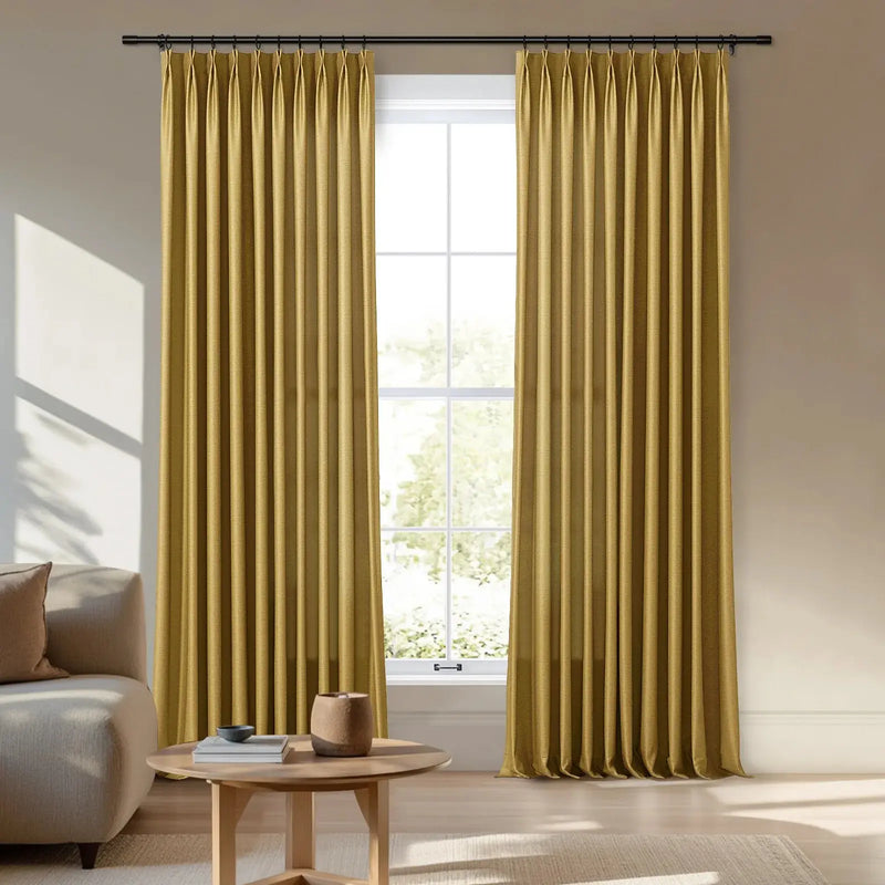 Floor-length golden pinch pleat curtains framing a window in a sunlit living room with a beige sofa and a light wood coffee table holding books and a pot